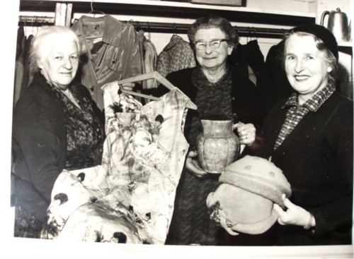 Three women in the 1960s holding various items for sale for fundraising purposes